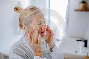 Beautiful senior woman in bathrobe, applying eye patches for puffiness while looking in the mirror photo