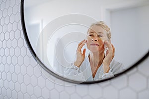 Beautiful senior woman in bathrobe, applying eye patches for puffiness while looking in the mirror photo
