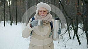 Beautiful senior woman drinking tea from thermos during enjoyable walk in winter park