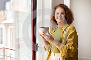 Beautiful senior woman enjoying cup of coffee while standing by the window