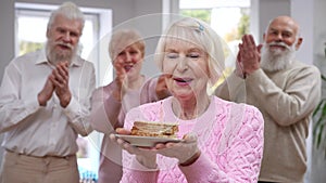 Beautiful senior woman making wish with closed eyes blowing out candle on birthday cake piece. Blurred old friends