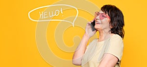 Beautiful Senior woman in pink neon sunglasses talking by the smartphone over yellow background