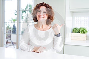 Beautiful senior woman wearing white sweater at home smiling with happy face looking and pointing to the side with thumb up