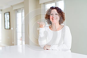 Beautiful senior woman wearing white sweater at home smiling with happy face looking and pointing to the side with thumb up