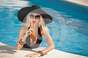 Beautiful sensual blonde with fashionable sunglasses relaxing in the pool with a juice. Attractive long hair woman in black