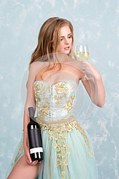 Beautiful sensual blonde woman in gorgeous long dress holding glass of white wine and bottle. Young girl celebrating