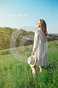 Beautiful sensual brunette in dress and hat standing with eyes closed in nature in bright back lit