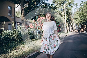 Beautiful sensual brunette young woman in white blouse and skirt with flowers walk on the street close to red roses bush