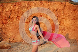 Beautiful sensual slim girl model in bikini and fashion sunglasses with blowing scarf on the beach at sunset.