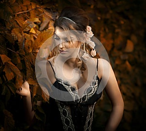 Beautiful sensual woman with roses in hair posing near a wall of green leaves. Young female in black elegant dress daydreaming