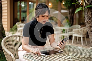 A beautiful serious Asian woman sits at an outdoor table of a cafe using her phone and tablet