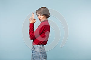 Beautiful serious girl with dark short hair in red sweater and jeans showing gun gesture with hands while thoughtfully