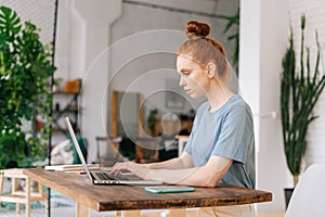 Beautiful serious young redhead woman is working on modern laptop computer at the table