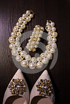Beautiful set of women`s wedding accessories. Beige shoes with shiny stones on them, pearl necklace and a bracelet on the wooden b