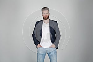 A beautiful sexy bearded man in a jacket holds his hands on his jeans and looks at the camera. he stands in front of the white