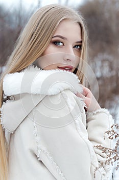 Beautiful blonde girl with long hair, full lips in a white coat walking in the winter woods