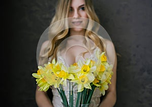 Beautiful blonde woman in beige lingerie with spring flowers bouquet of daffodils. Selective fokus in flowers