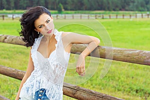 Beautiful brunette girl with red lips in the white shirt in denim shorts standing near the horse paddock