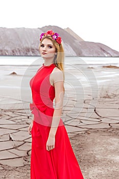 Beautiful cute girl with long blond hair in a long red evening dress with a wreath of roses and orchids in her hair standing