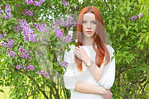 Beautiful cute red-haired girl in a white dress standing near bushes with lilac