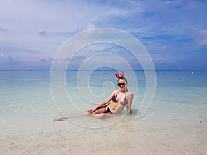 beautiful sexy fit woman in bikini and sunglasses sitting in blue water on beach. Female wearing christmas reindeer headband