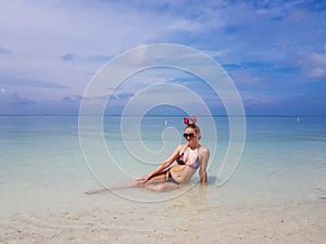 beautiful sexy fit woman in bikini and sunglasses sitting in blue water on beach. Female wearing christmas reindeer headband