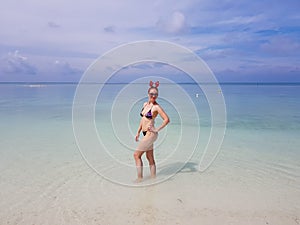 beautiful sexy fit woman in bikini and sunglasses standing in blue water on beach. Female wearing christmas reindeer headband