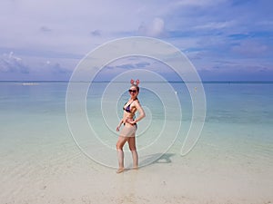 beautiful sexy fit woman in bikini and sunglasses standing in blue water on beach. Female wearing christmas reindeer headband