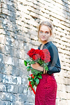 Beautiful and girl with a bouquet of red roses stands on the background of an old brick wall.