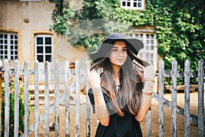 Beautiful girl brunette with brown eyes in a black dress and a black hat with big brims against the backdrop of a wooden fenc