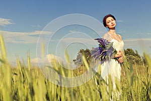 Beautiful girl with dark hair in white sundress with a bouquet of flowers lupine walks on the field with rye on a sun