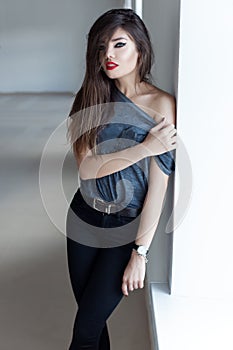 Beautiful girl with long dark hair rocker style with red lipstick on gubyh standing near a window in a black leather jacket