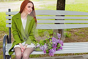 Beautiful girl with long hair in ryzhymi green coat sitting on a park bench with a bouquet of lilac in the hands of