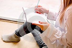 Beautiful sexy girl sits on a windowsill with a notebook and thinks. Details close up. A lonely woman in a man`s shirt reads a bo