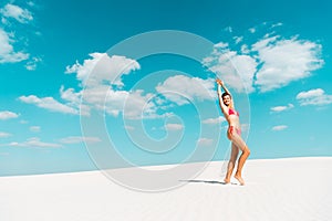 Beautiful sexy girl in swimsuit with hands in air on sandy beach with blue sky and clouds