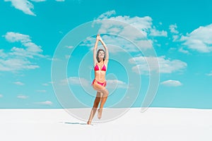 Beautiful sexy girl in swimsuit jumping with hands in air on sandy beach with blue sky and clouds
