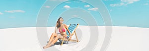 Beautiful sexy girl in swimsuit and sunglasses sitting in deck chair on sandy beach with blue sky and clouds, panoramic