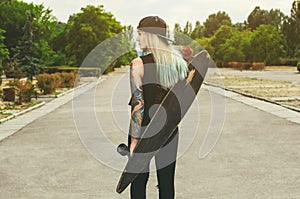 Beautiful, hipster blonde with blue hair in tattoo stands with a longboard it is turned back on the street.