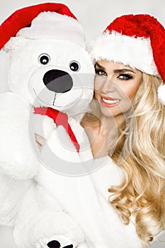 Beautiful sexy, smiling blonde model dressed in a Santa Claus hat, holding a teddy bear. Beauty sensual girl for Christmas