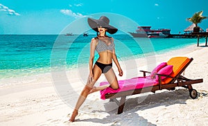 Beautiful sexy tanned woman bikini model at Maldives tropical sand beach. Young glamour girl in swimsuit and hat on amazing