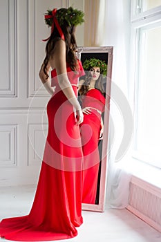 Beautiful woman in elegant long evening red dress standing in the mirror next to the window with a Christmas wreath on her