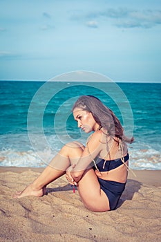 Beautiful woman with tan skin in a black swimsuit sitting on the sand of the beach. Female hug her knees near sea.