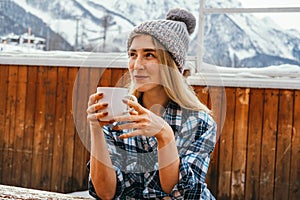 Beautiful sexy woman wearing knitting hat and holding hot drink mug outdoor on a wooden terrace in winter mountains