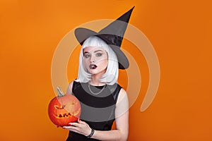 Beautiful sexy woman in witch hat and costume holding carved pumpkin on orange background.