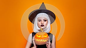 Beautiful sexy woman in witch hat and costume holding carved pumpkin on orange background.