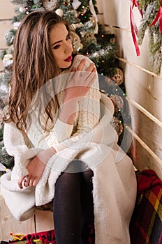 Beautiful sexy woman with Xmas tree in background sitting on elegant chair in cozy scenery. Portrait of girl posing
