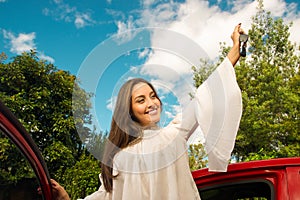 Beautiful young woman wearing a white blouse coming out of her red car and holding a keys while she is smiling