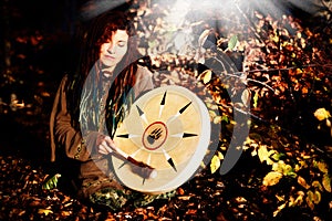 Beautiful shamanic girl playing on shaman frame drum on background with leaves and flowers.