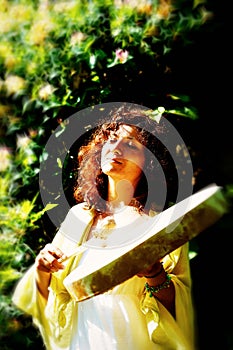 Beautiful shamanic girl playing on shaman frame drum on background with leaves and flowers.