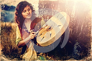 Beautiful shamanic girl playing on shaman frame drum in the nature, old photo effect.